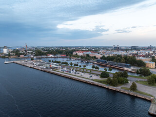 Obraz premium Tallinn waterfront with a marina, docked boats, a paved promenade, parking spaces, colorful buildings, a tall chimney, and a cloudy sky.
