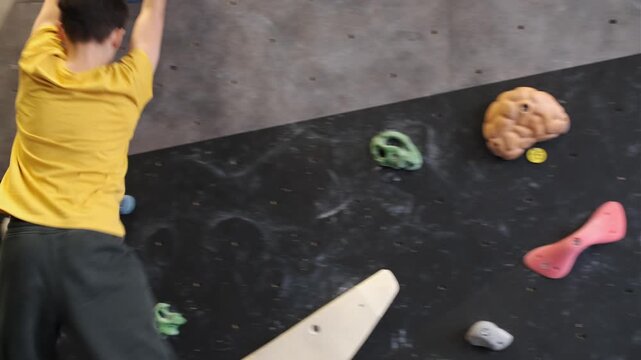 Side view of a teenage boy climbing on an indoor bouldering wall while gripping a large blue sloper volume.