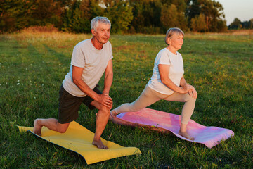 Active and fit elderly couple doing a lunge stretch on yoga mats in a field at sunset.