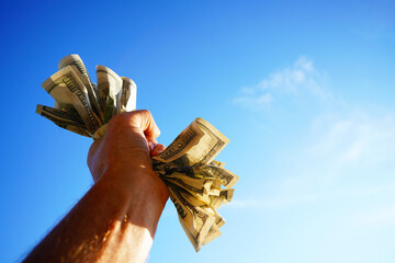 Dollars in hand on blue sky background. Crumpled pile of dollar bills in male hand on blue sky with space for text. Person clutching a handful of crumpled dollar bills tightly. Crumpled Money in hand
