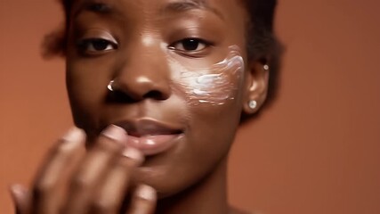 Serene Young African American Woman Applying Cream to Her Face in Close Up with a Natural Expression Against a Warm Brown Background for Beauty and Skincare Product Advertisement and Promotion