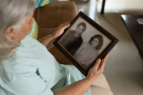 A rich senior lonely elder woman revisiting memories by watching old family picture frame while sitting alone on sofa on living room - Powered by Adobe