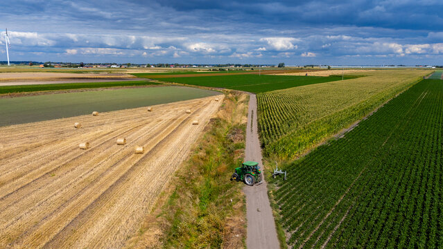 Aerial drone view of farmland, hay bales, green tractor on rural dirt road, agricultural landscape with wind turbine and dramatic clouds on horizon.