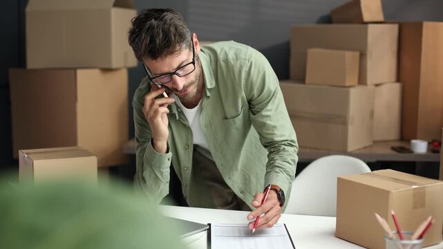 A man is talking on his cell phone while sitting at a desk with a stack of cardb