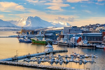 Snow-covered Troms harbor with docked boats and ships, nestled against a backdrop of snow-capped mountains and colorful buildings. A serene winter scene capturing the essence of Norwegian coastal life