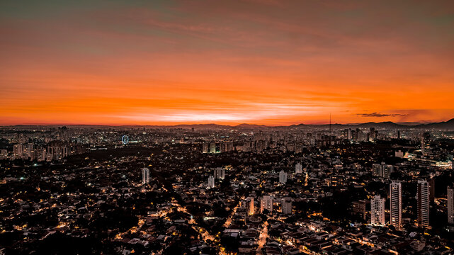 Aerial drone view of S&atilde;o Paulo sunset over Vila Madalena, Vila Beatriz and Alto de Pinheiros neighborhoods, with city lights, skyline silhouette, and vibrant orange sky captured by DJI Mavic 3