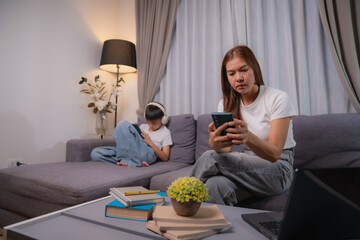 Asian family, mother using smartphone for social media or working and her son playing game on tablet, sitting on sofa in living room at night, concept of social problem and family issues