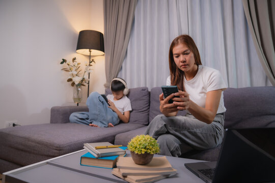 Asian family, mother using smartphone for social media or working and her son playing game on tablet, sitting on sofa in living room at night, concept of social problem and family issues