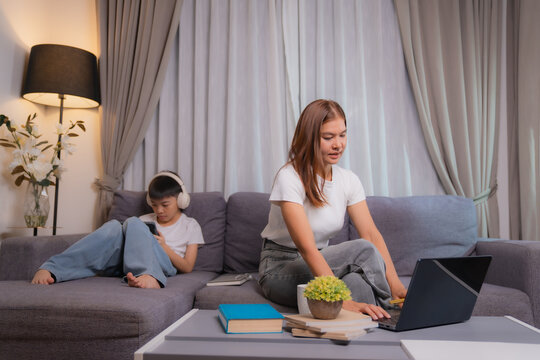 Asian businesswoman teleworking on laptop while her son is using smartphone and listening music with headphones, sitting on sofa in living room at home