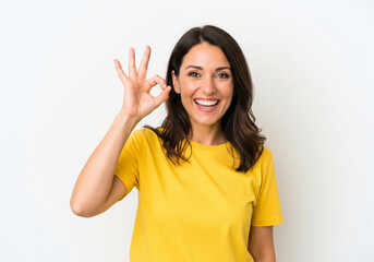 Fototapeta premium Smiling woman in yellow shirt makes an OK hand gesture against a white background, expressing approval and positivity.