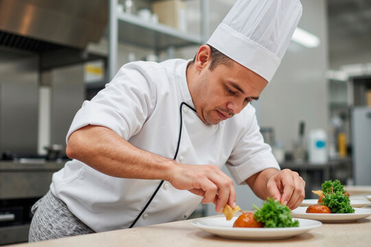 Middle aged Caucasian man chef preparing gourmet dish in professional kitchen, carefully arranging food on plate with focused expression, wearing traditional chef uniform and hat
