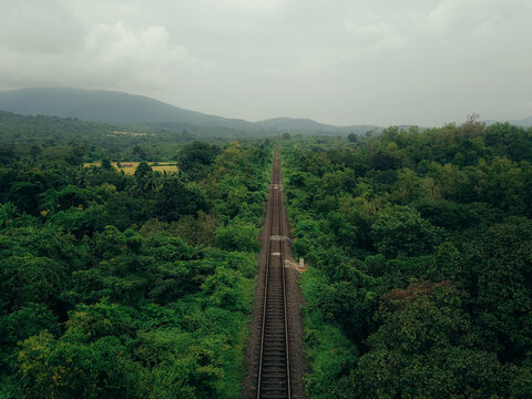Aerial view of a solitary railway track slicing through a dense, verdant forest under a somber sky, Neturlim, Goa, India.