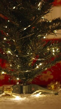 Vertical video. Close Up of Base of Christmas Tree Wrapped in Lights with Ornaments and Woman in Red Pajamas Sitting in Background