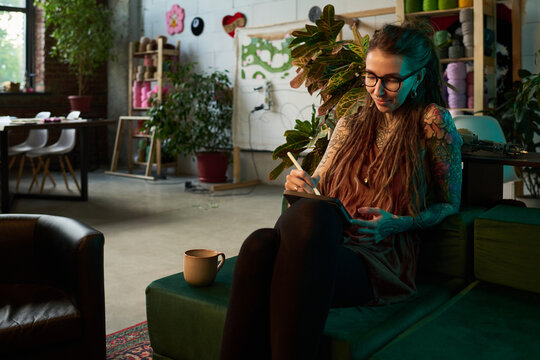 Young adult Caucasian woman with long hair and tattoos sitting on bench sketching carpet design in workshop surrounded by plants and materials, focused on creative process