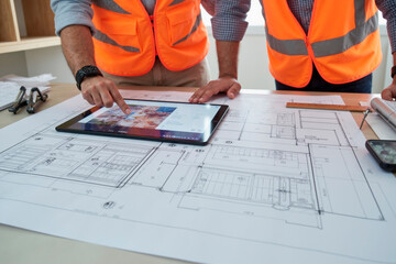 Two middle aged Caucasian men wearing safety vests reviewing digital building plans on tablet while standing over architectural blueprints on desk, collaborating on construction project