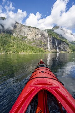 Kayak trip on the Geirangerfjord with waterfall The Seven Sisters, tip of a red kayak in the fjord, atmospheric fjord landscape with reflection, first person view, near Geiranger, M&oslash;re og Romsdal, Norway