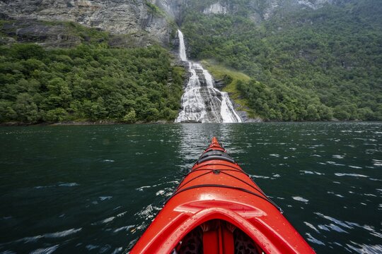 Kayak trip on the Geirangerfjord at the Freier waterfall, tip of a red kayak in the fjord, atmospheric fjord landscape, first person view, near Geiranger, M&oslash;re og Romsdal, Norway