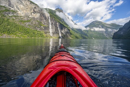 Kayak trip on the Geirangerfjord with waterfall The Seven Sisters, tip of a red kayak in the fjord, atmospheric fjord landscape with reflection, first person view, near Geiranger, M&oslash;re og Romsdal, Norway