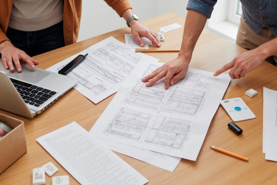 Caucasian young adult man and Caucasian young adult woman collaborating on architectural project, reviewing blueprints and technical drawings on desk with laptop and measuring tools