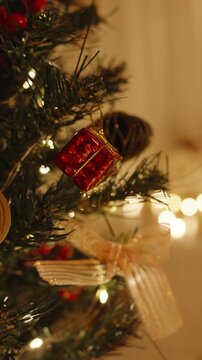 Vertical video. Close Up of Hand Placing Red Gift Box Ornament on Tree with Glowing Lights and Blurred Background of Cozy Room