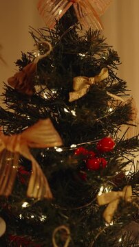 Vertical video. Woman in Santa Hat Holding Ornament and Preparing to Decorate Mini Christmas Tree in Cozy Room with Warm Lights