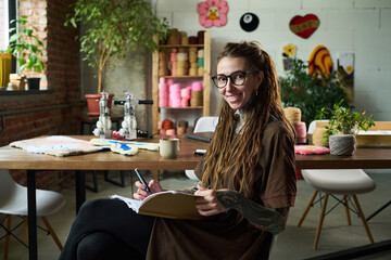 Portrait of young adult Caucasian woman with glasses and tattoos sitting at table in carpet workshop smiling at camera, while sketching designs with yarn and textile samples visible