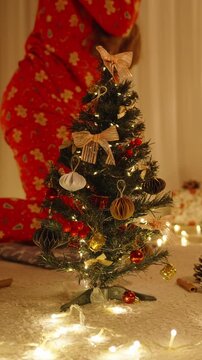 Vertical video. Woman in Santa Hat Sitting Near Decorated Mini Christmas Tree in Cozy Room with Gift Boxes and Glowing String Lights