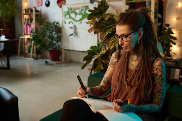 Caucasian young adult woman with tattoos and dreadlocks sitting in carpet workshop sketching designs in notebook, surrounded by plants and creative workspace elements