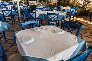 Authentic Greek taverna terrace with classic blue and white table settings, stone flooring, and traditional wooden chairs awaiting seaside diners.