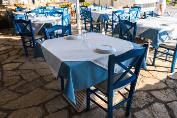 Authentic Greek taverna terrace with classic blue and white table settings, stone flooring, and traditional wooden chairs awaiting seaside diners.
