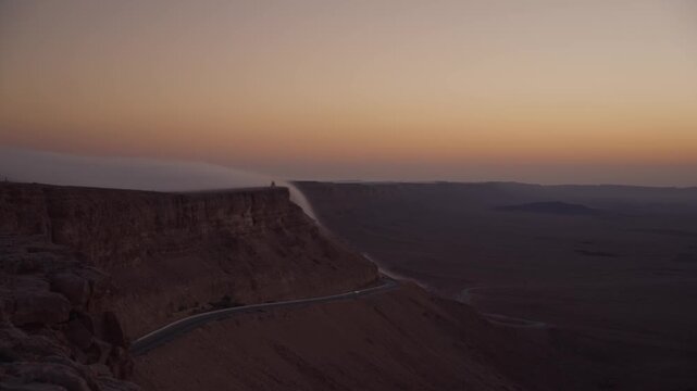 Stunning early morning mountain cliff as sun is rising over the Ramon Crater in the Negev region desert in Israel, with fog rolling as a cloud waterfall phenomenon