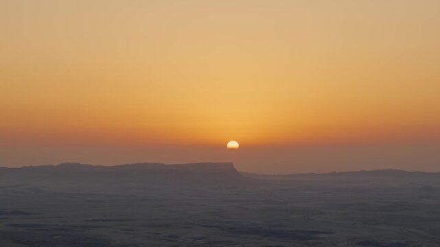 Majestic view of the sun rising slowly behind a desert mountain range, casting a warm golden glow across the hazy sky and silhouetting the rugged landscape in Ramon Crater in Israel Negev region