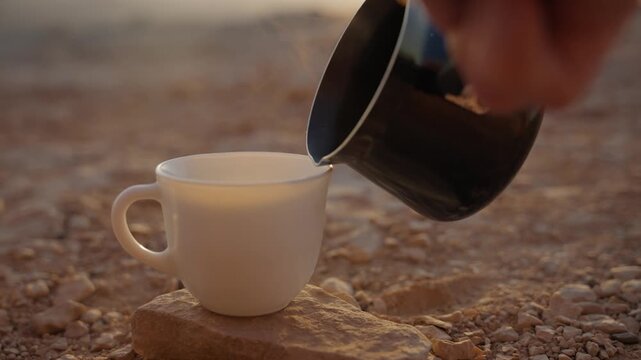 Close up of briki or cezve pot pouring tea into small ceramic cup in desert. Traditional hot drink preparation in Middle East, Hiker or Beduin hospitality. Camping outdoors. Hot drink poured into mug