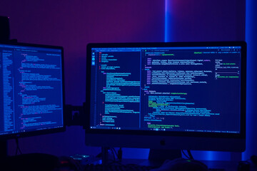 Dual computer monitors displaying programming code in dark room, screens showing lines of software development scripts and code editor interface, technology workspace setting