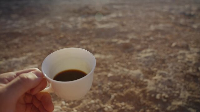 Hand holding coffee cup in desert background in early morning trip or hiking. Camping outdoors in Middle East desert. Drinking hot black coffee traditional arabic ground cafe. POV male hand holding