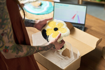 Young adult Caucasian woman with tattooed arms holding handmade floral tufted rug, placing finished piece into cardboard box in carpet workshop with laptop visible in background