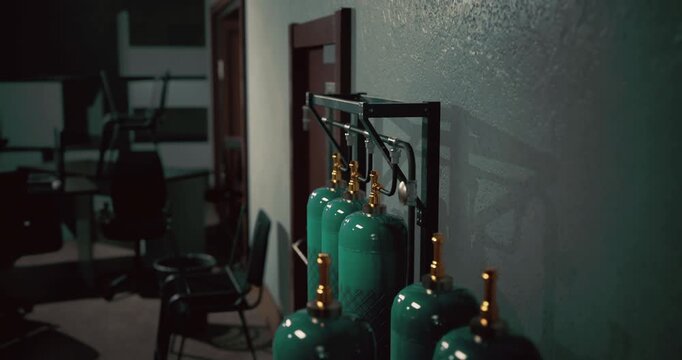A row of green gas cylinders stands against a textured wall in a shadowy setting. Soft light highlights the bright valves, adding a touch of intrigue.