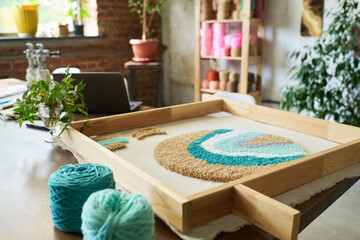 Wooden frame holding unfinished abstract carpet design resting on worktable in carpet workshop with turquoise yarn balls and laptop nearby, shelves filled with colorful yarn in background