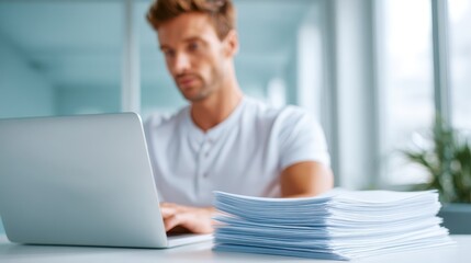 Fototapeta premium Man working at clean minimalistic desk with laptop and large stack of documents, modern office environment, focused and productive atmosphere