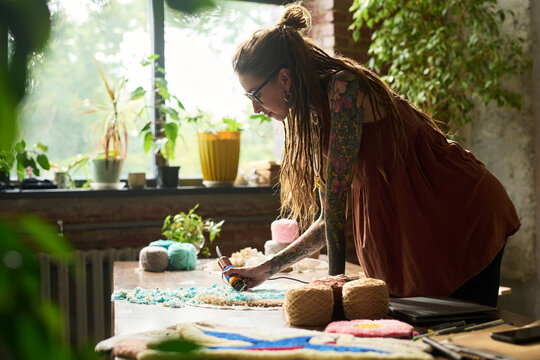 Young adult Caucasian woman with long dreadlocks and tattooed arms working on handmade carpet using tufting tool in carpet workshop, natural light streaming through window, yarn balls on table