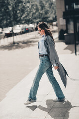 Fototapeta premium Stylish brunette woman in a grey trench coat walking confidently in an urban street setting under sunlight outdoors, vertical portrait