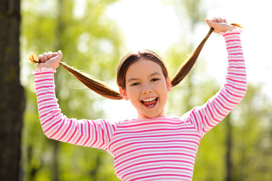 Smiling young girl outdoors pulling her brown pigtails, wearing a pink striped shirt, enjoying a lively spring day - Powered by Adobe