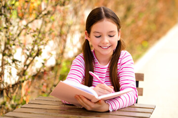 Smiling young girl writing in notebook outdoors on sunny day, enjoying casual leisure time in striped shirt during spring.