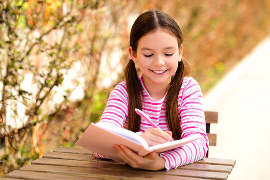 Smiling young girl writing in notebook outdoors on sunny day, enjoying casual leisure time in striped shirt during spring. - Powered by Adobe
