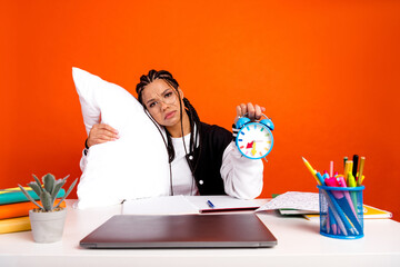 Teen girl holding alarm clock and pillow in a classroom setting with colorful school desk supplies and orange background