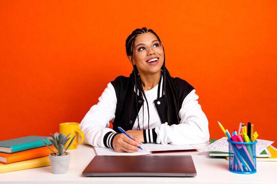 Smiling young female student with braided hair at desk with notebook, laptop, and book on bright orange background