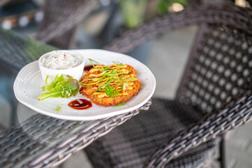 Breaded meat cutlet with sauce, white rice, greens, and ketchup served on a plate atop a glass table in a café setting with rattan chair in background. Horizontal, daylight.
