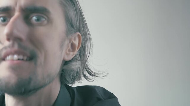 Close-Up of a Quirky Man with Medium-Length Hair and Beard Transitioning from a Serious Expression to a Joyful, Playful Smile with a Fun Look in a Soft Background