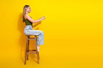 Smiling young woman sitting on a wooden stool wearing casual style with khaki top and jeans against...