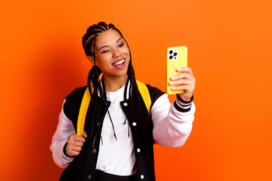 Confident young woman with braids taking a selfie on an orange background, showing happiness and charisma